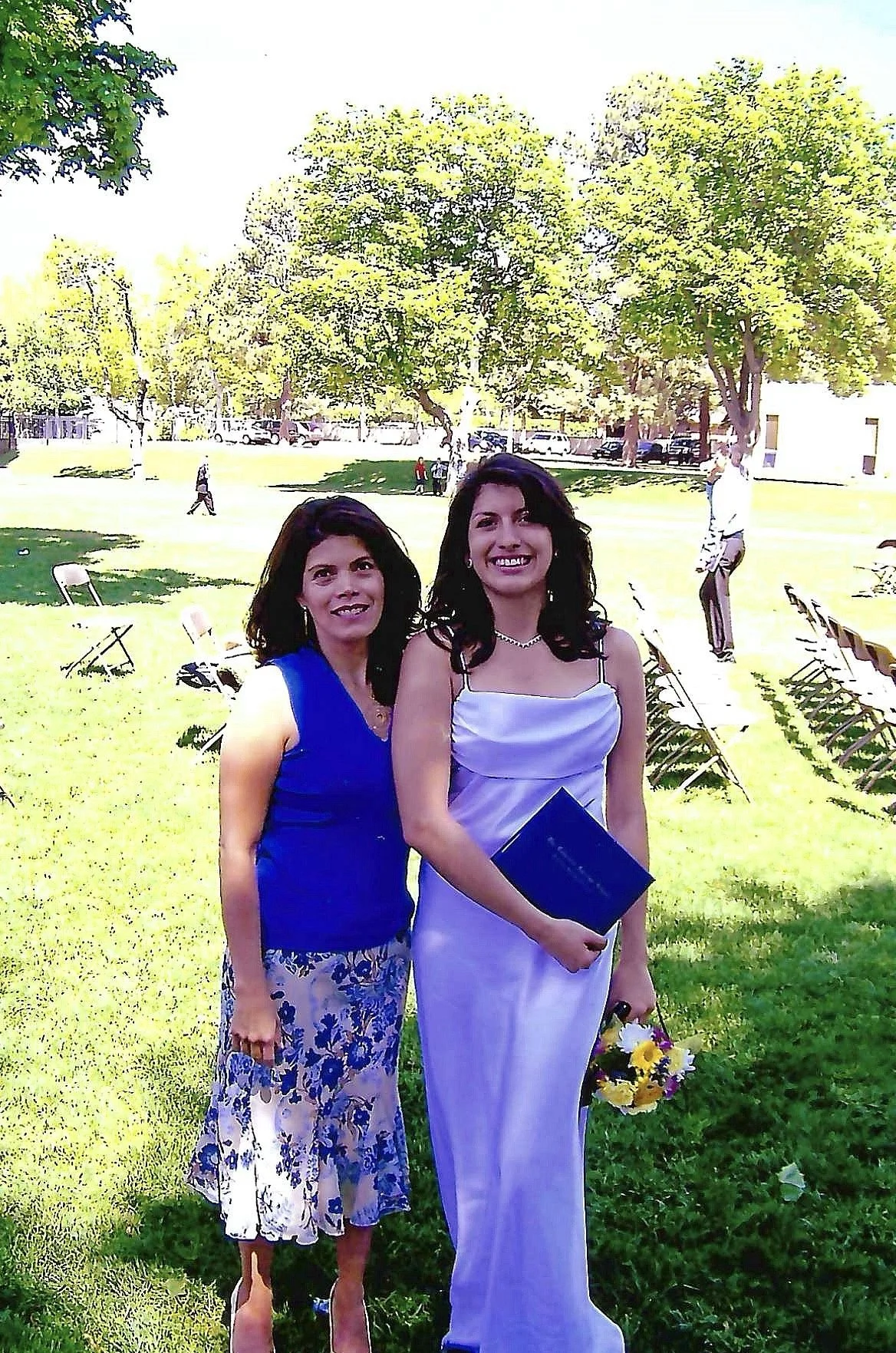 Two women standing outside on a sunny day, one in a white dress holding a bouquet of flowers, and the other in a blue top with a floral skirt.