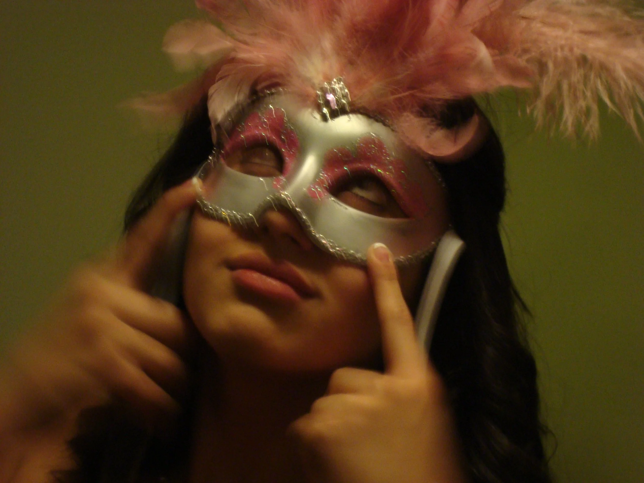 Person wearing a silver and pink masquerade mask with a feathered pink accessory on top, touching the mask's side with their finger.