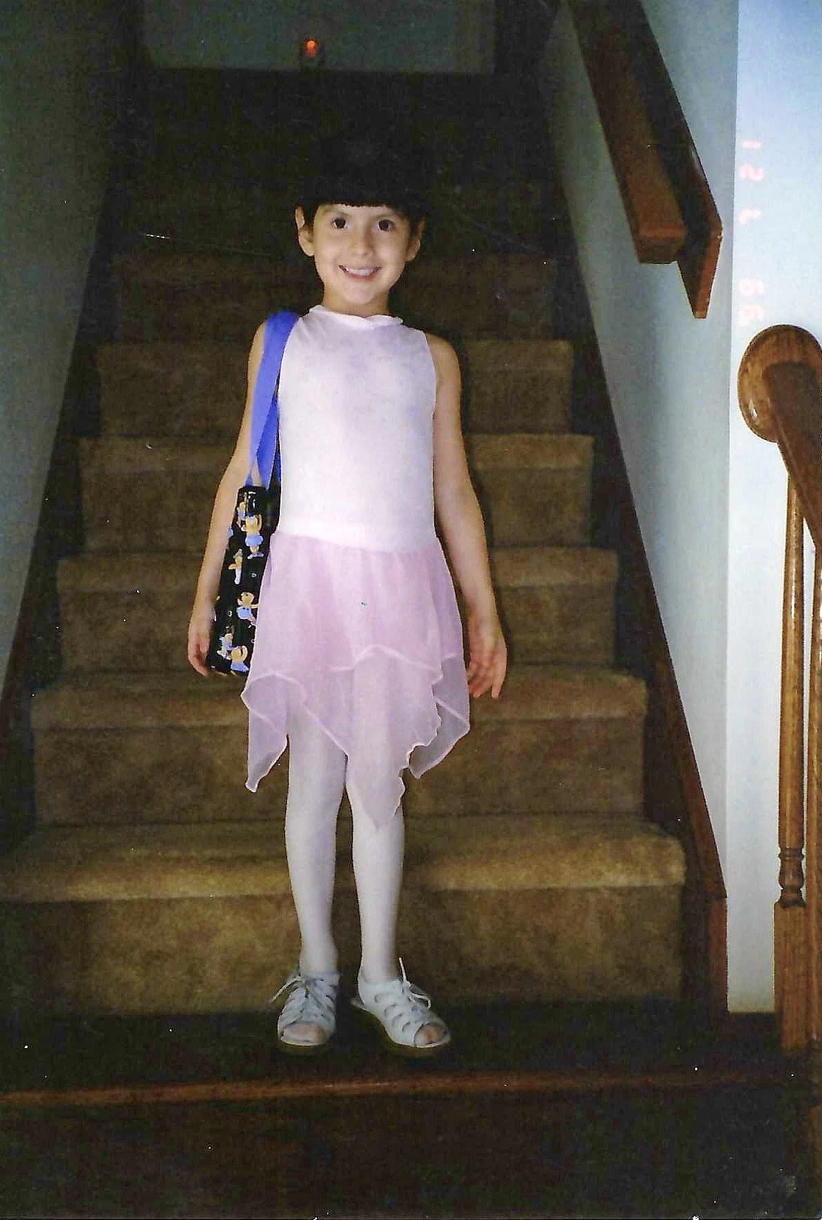 Young girl standing on a staircase wearing a ballet costume with a pink and white dress, white tights, and ballet shoes, smiling at the camera.
