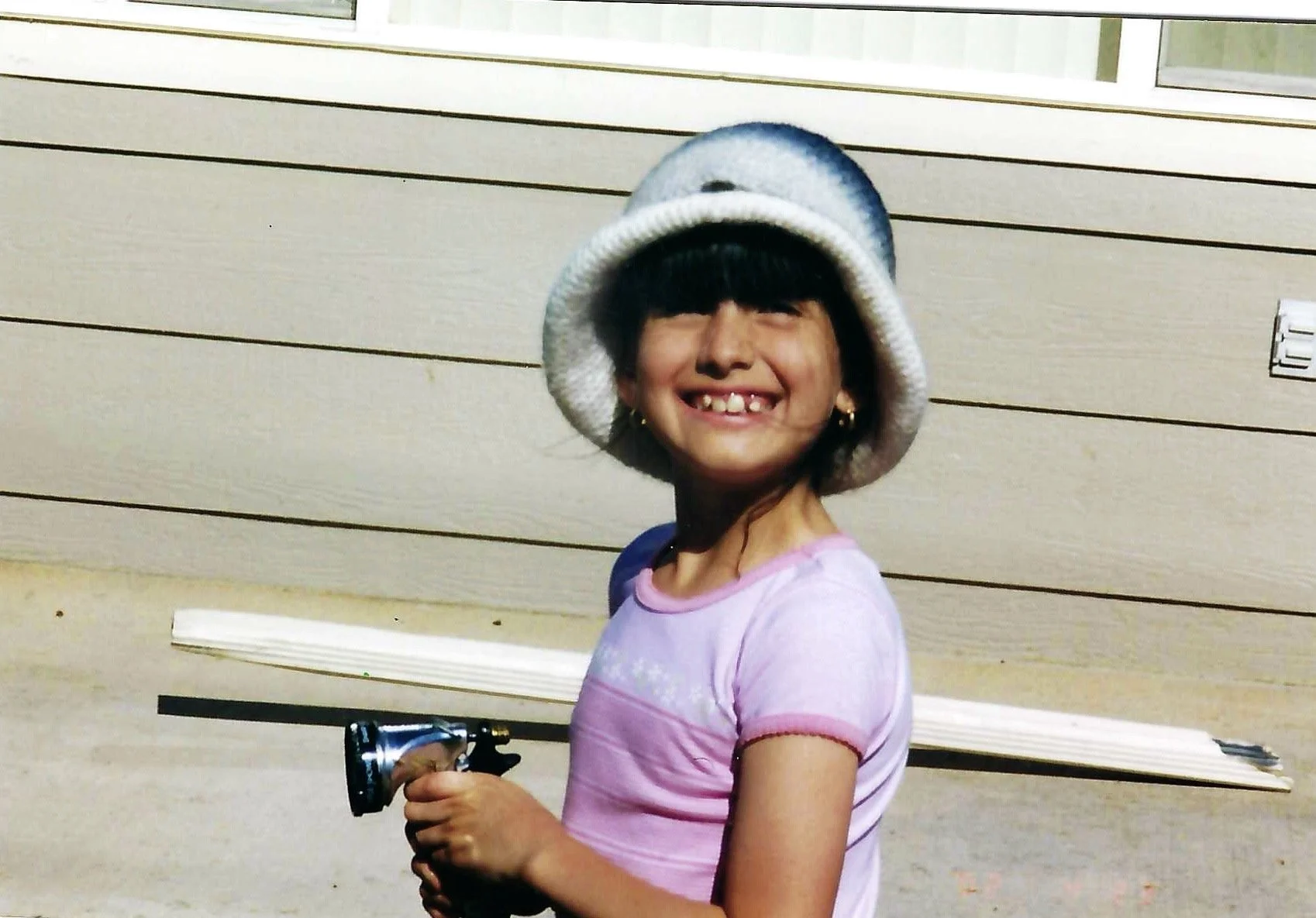 A smiling young girl with missing front teeth, wearing a gray knit hat with a white brim, a pink tie-dye shirt, gold earrings, and holding a toy gun, standing outdoors in front of a beige wall with horizontal siding.