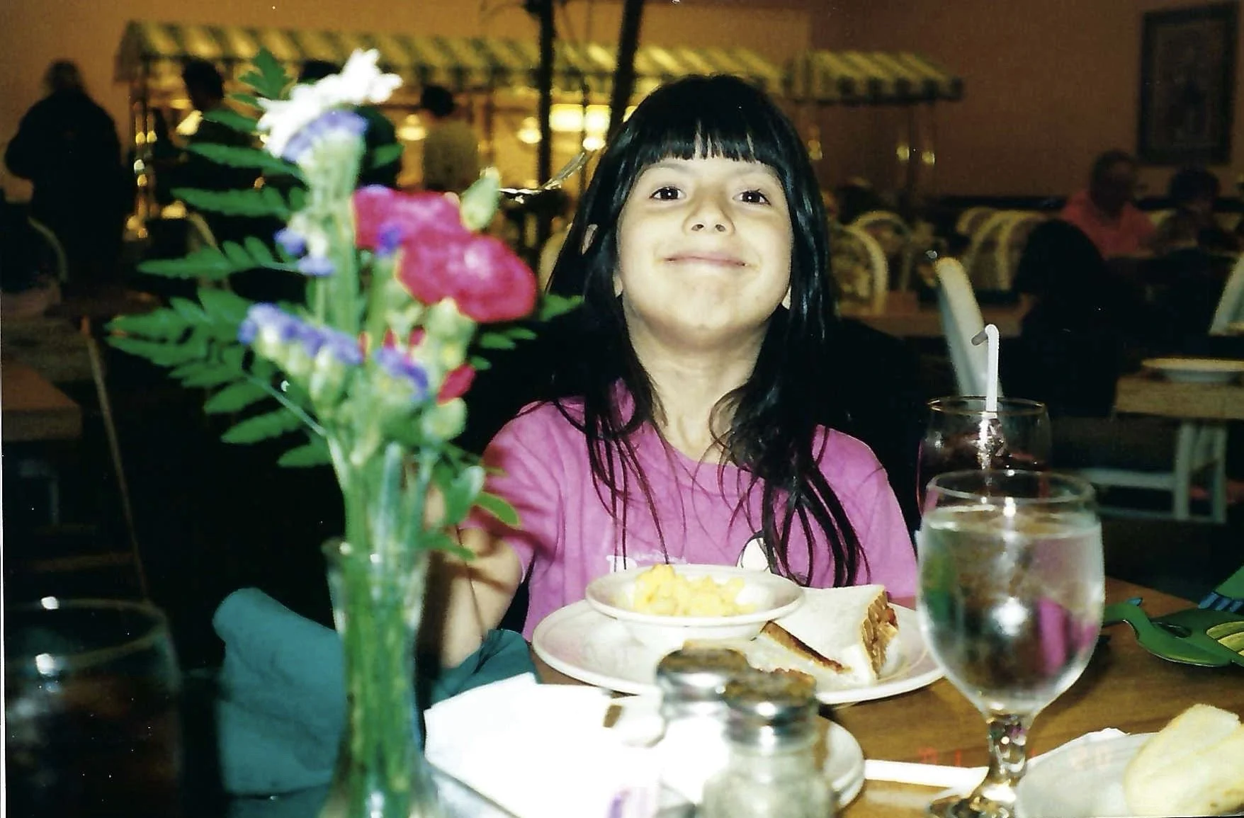 Child with long dark hair and a pink shirt smiling at a restaurant table with food, flowers, and drinks in the foreground.