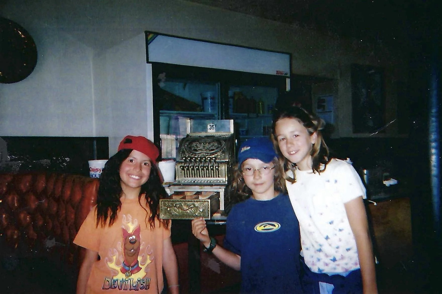 Three young girls smiling and posing for a photo inside a restaurant. One girl is wearing an orange T-shirt with a cartoon character and a red cap, the second girl has glasses and is wearing a blue cap, and the third girl is dressed in a white shirt 