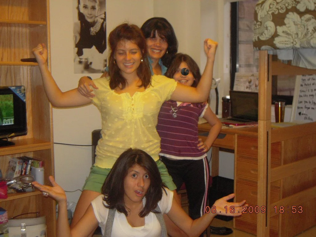 Four girls in a room posing playfully. One girl in a yellow top and green shorts is standing with her arms flexed. Another girl in a purple striped shirt and sunglasses is standing behind her. A third girl appears to be sitting or crouching in front 