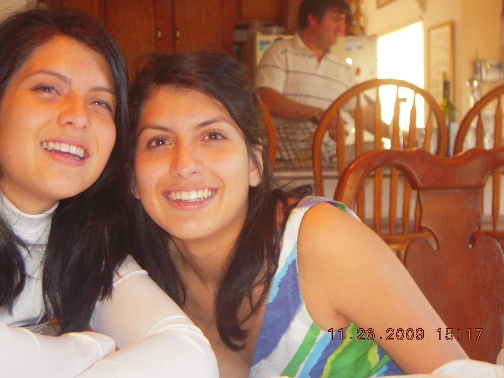 Two young women smiling at a table with wooden chairs, with a man in a striped shirt in the background in a kitchen.