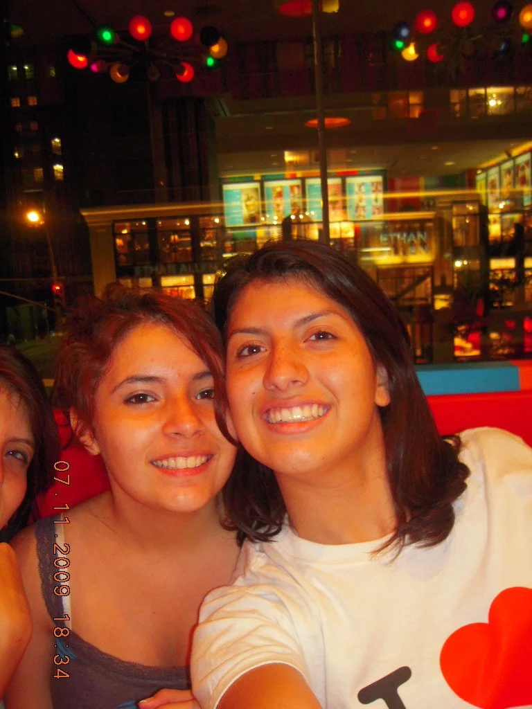 Two women smiling and taking a selfie in a restaurant or cafe with colorful lights and a view of a city street at night in the background.