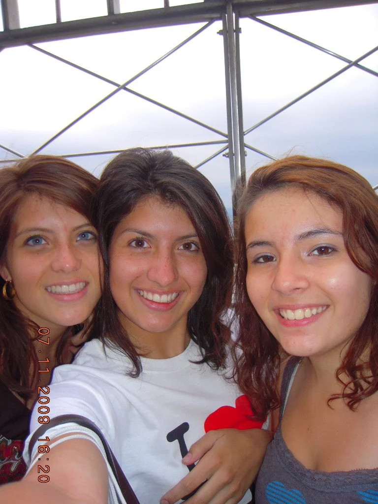 Three young women smiling and taking a selfie outdoors with a metal structure and cloudy sky in the background.