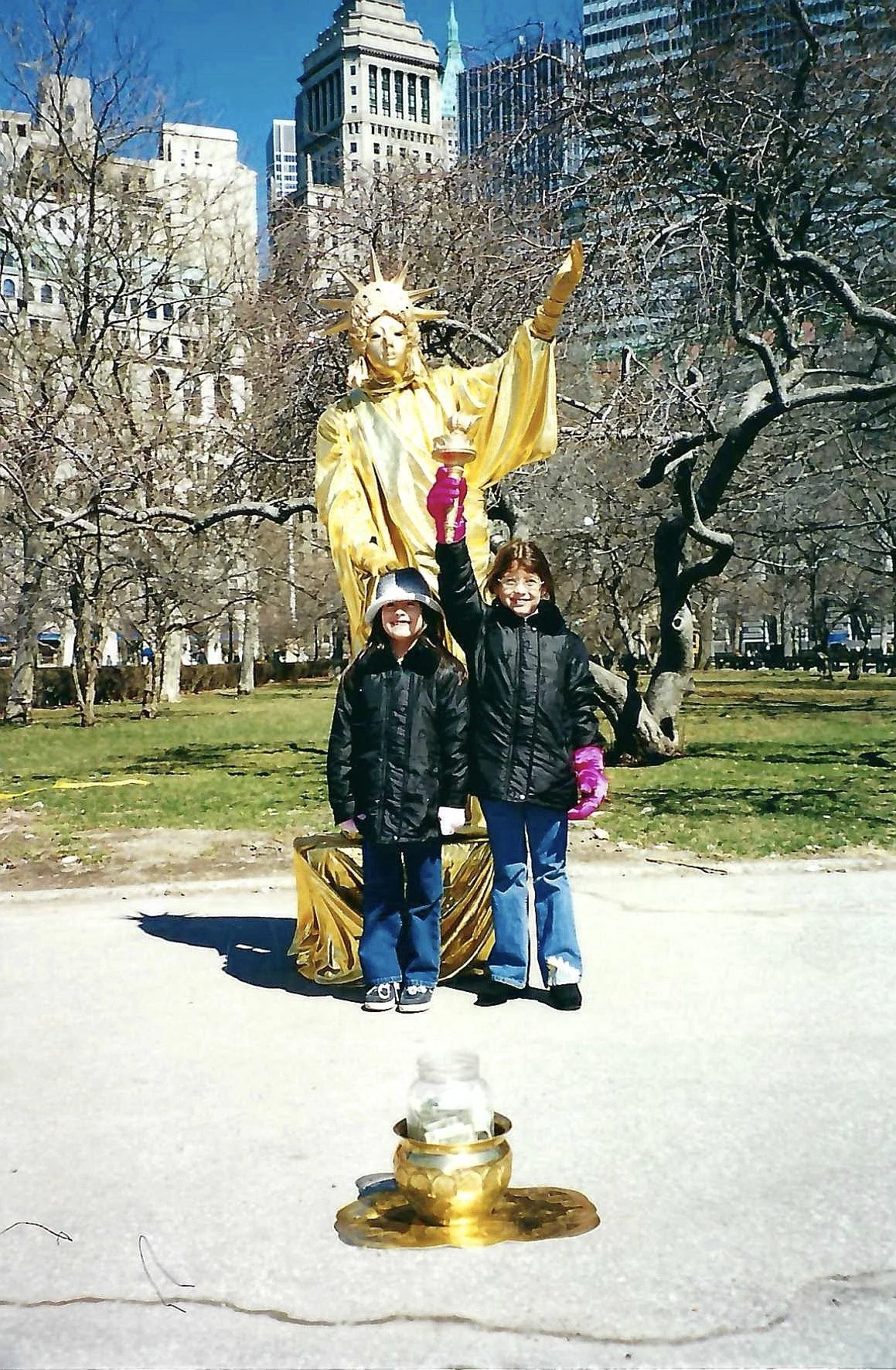 Two young girls standing outdoors in front of a golden Statue of Liberty sculpture, with bare trees and city buildings in the background. One girl is holding a small item up in the air, smiling, while the other girl stands beside her with a neutral e
