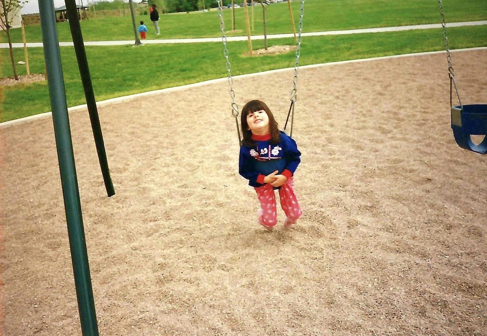 A young girl in pink pajama pants and a navy blue jacket is sitting on a swing in a park with green grass and trees, with other children and adults in the background.