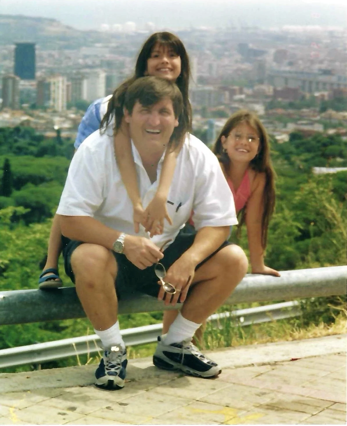 A man crouching on a roadside barrier with two young girls, one on his shoulders and the other sitting on the barrier, with a cityscape and green hill in the background.