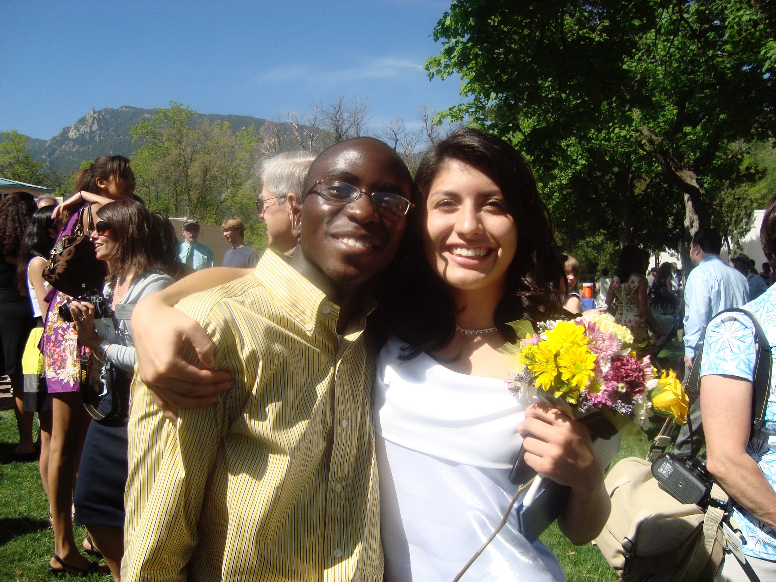 A group of people outdoors at a gathering, with two people in the foreground taking a selfie. The man is wearing glasses and a yellow striped shirt, and the woman is holding a bouquet of flowers and smiling. The background shows a sunny day with tree