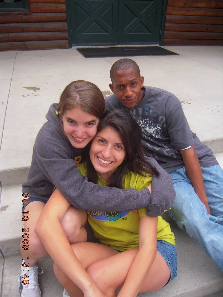 Three friends sitting on a step outside a wooden building, smiling and hugging each other.