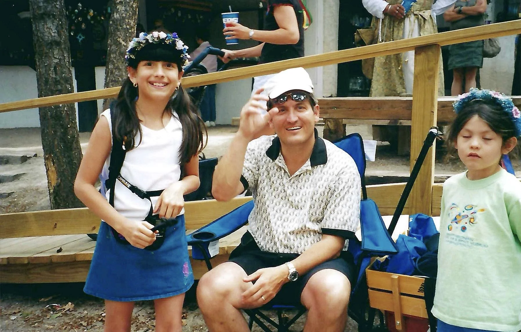 Three children and a man at an outdoor event, with the girl on the left smiling, the man in the middle wearing a white cap and waving, and the girl on the right with closed eyes, all seated or standing near a wooden fence.