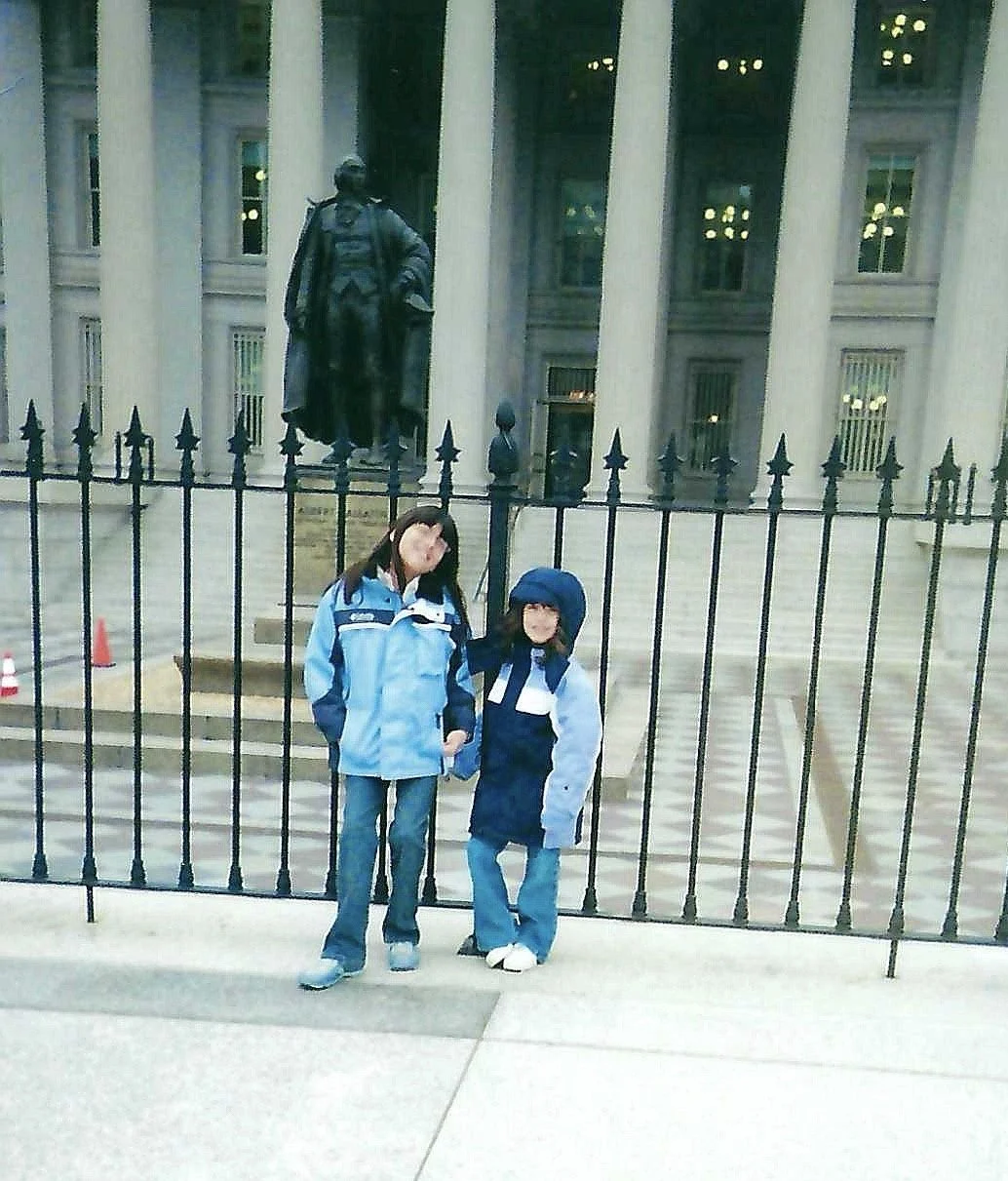 Two children standing in front of a black iron fence, with a large building and a statue in the background, during daytime.