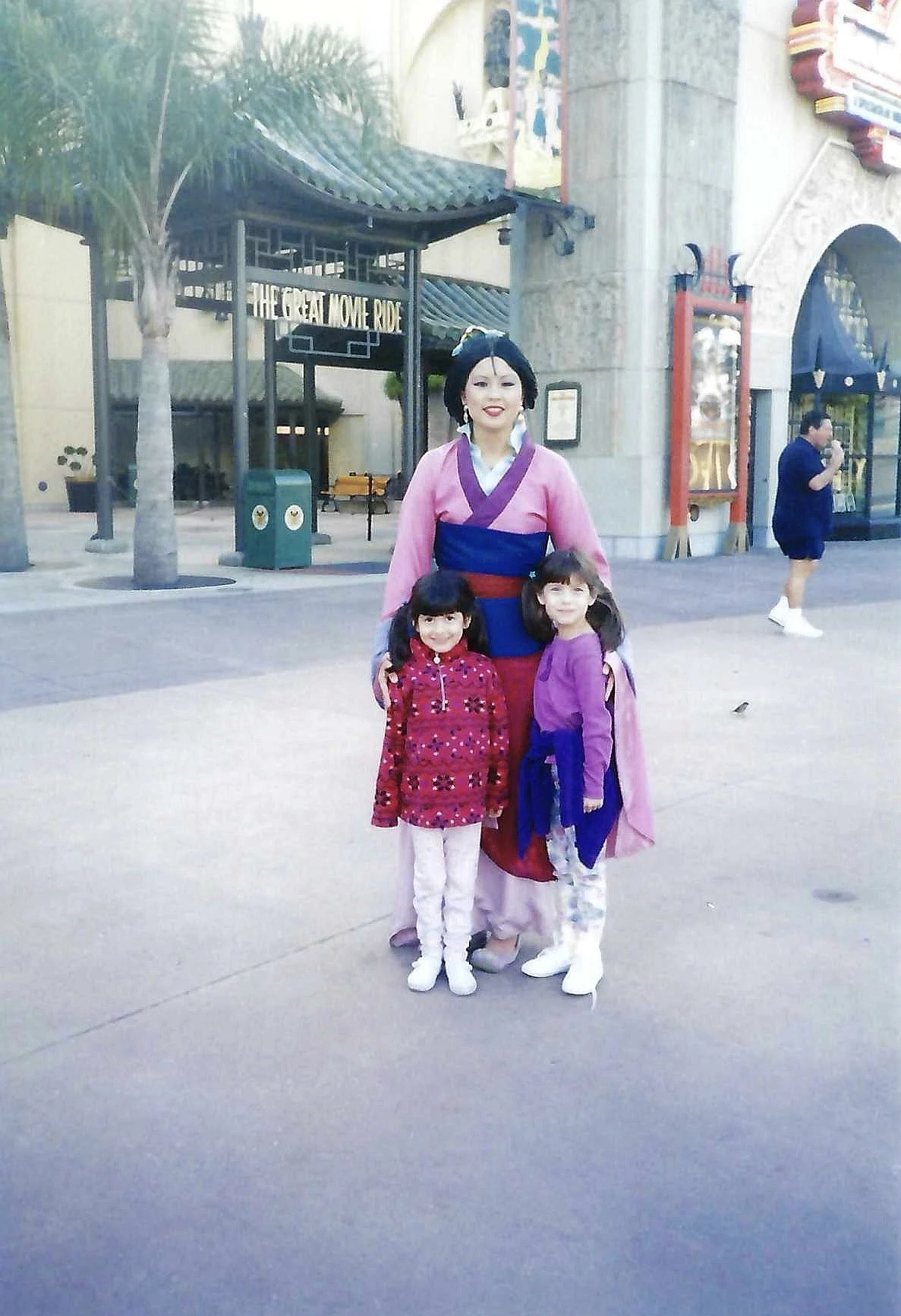 A woman dressed as Mulan from Disney's Mulan is standing with two young girls in front of an amusement park entrance, with a sign reading 'The Great Movie Ride' in the background.