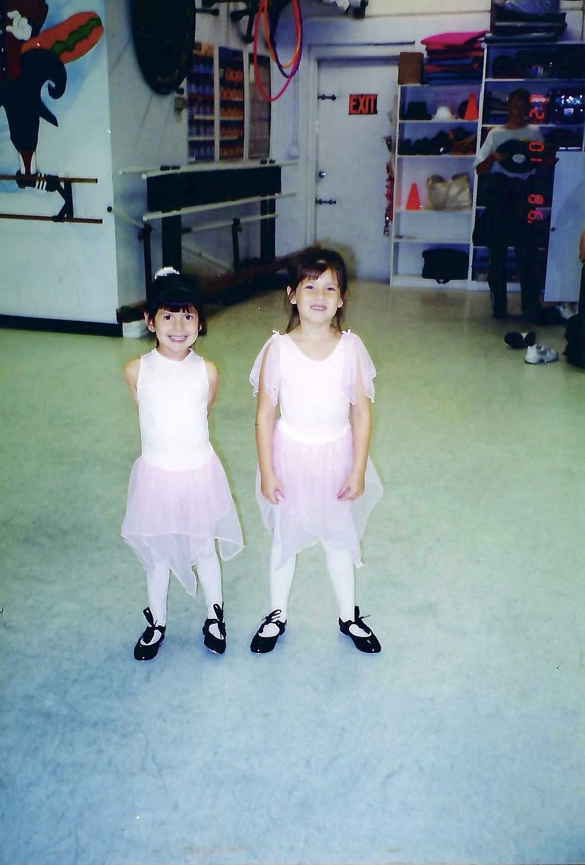 Two young girls standing in a dance studio, dressed in ballet costumes, smiling at the camera.