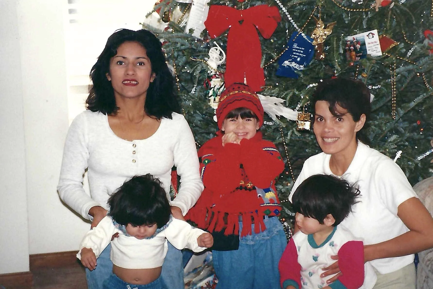 Family gathered around a decorated Christmas tree, smiling, with children and adults wearing festive holiday clothes.