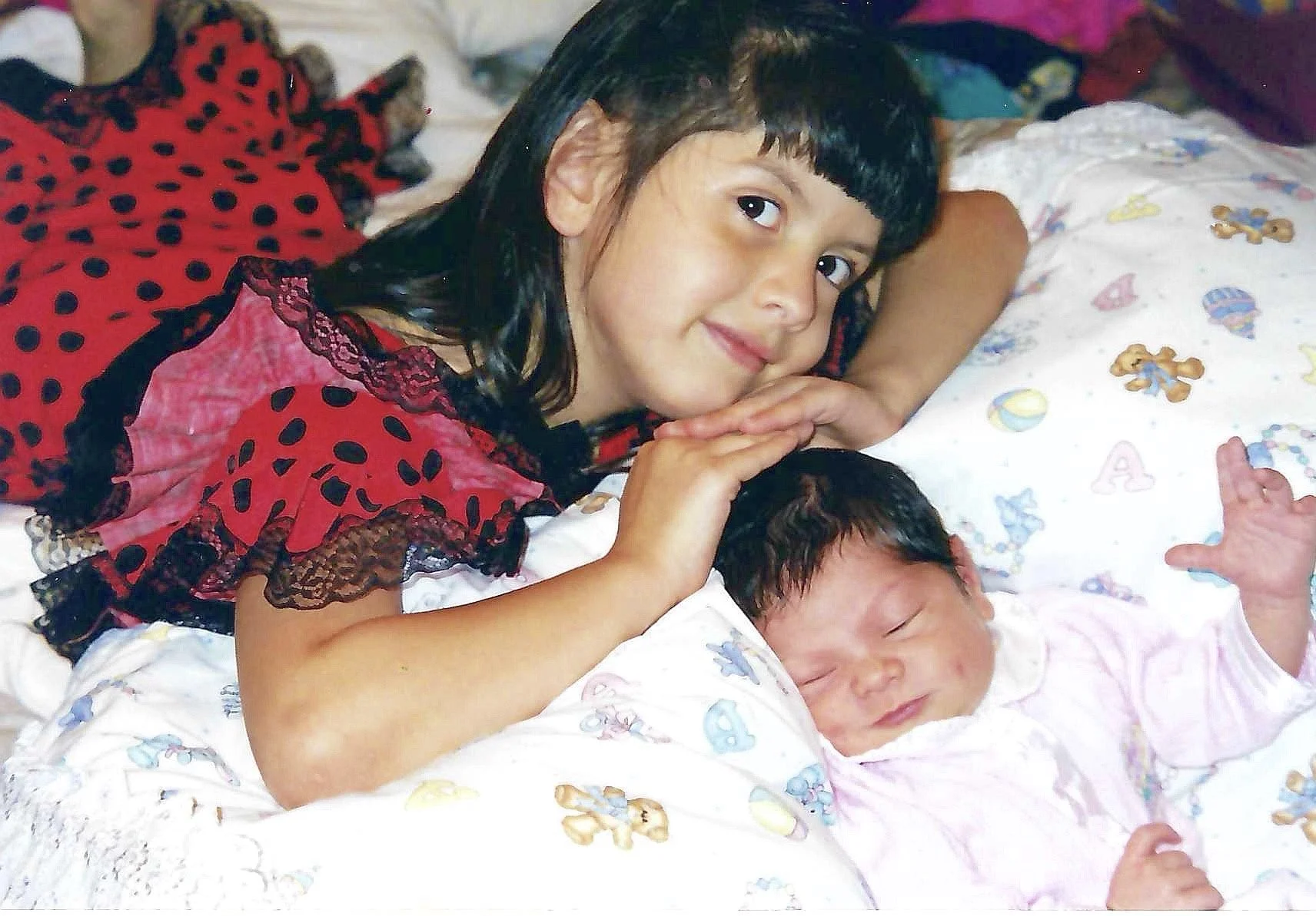 A girl with dark hair and a red dress with black polka dots, lying on her stomach on a bed with two sleeping babies, one with a pink striped shirt and the other in a white outfit, with colorful teddy bear and letter themed sheets.