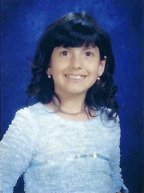 Young girl with dark hair and a bright smile, wearing a light-colored top and earrings, against a blue background.