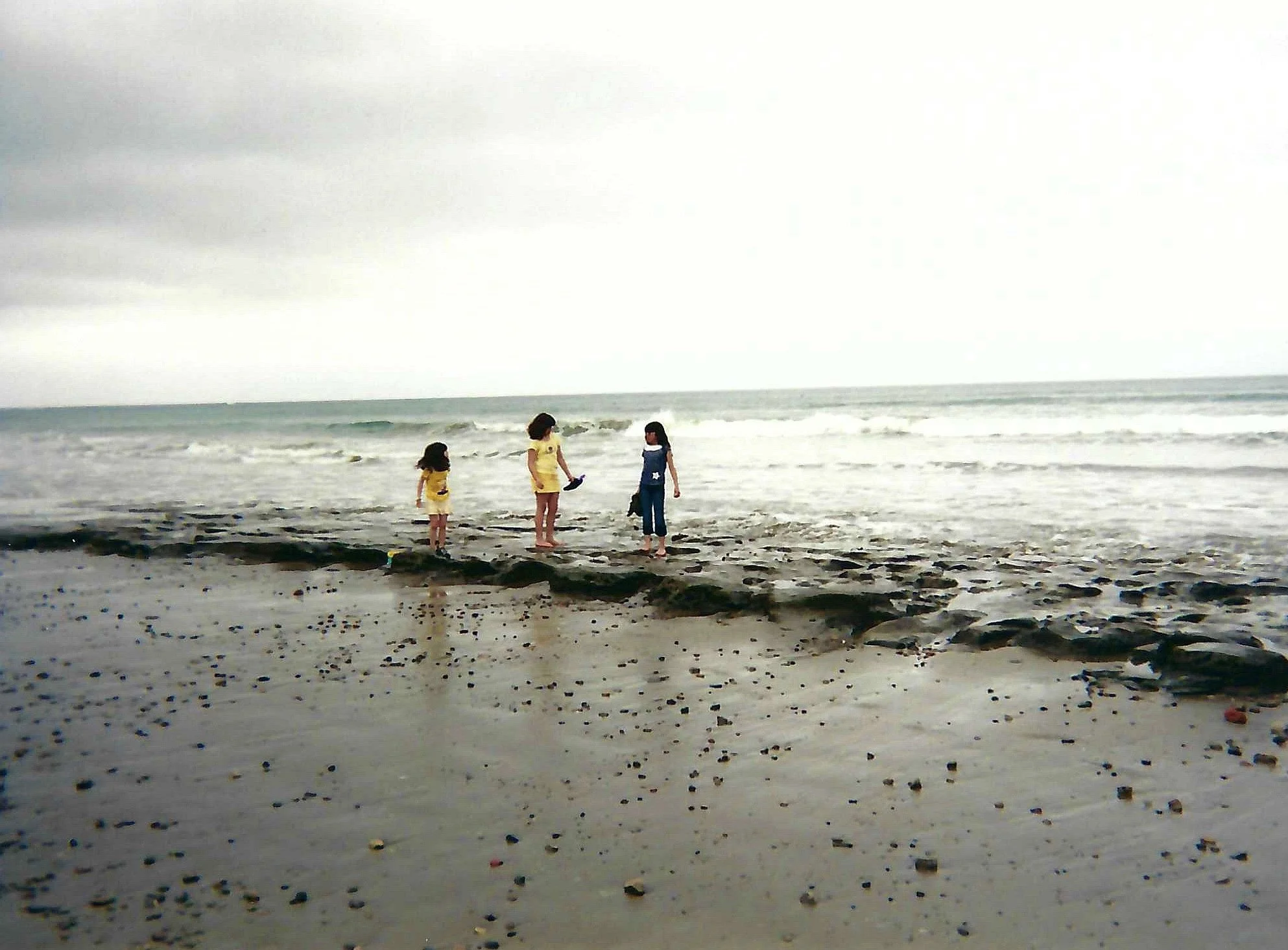Three children standing on a sandy beach near the water, looking out at the ocean, under a cloudy sky.