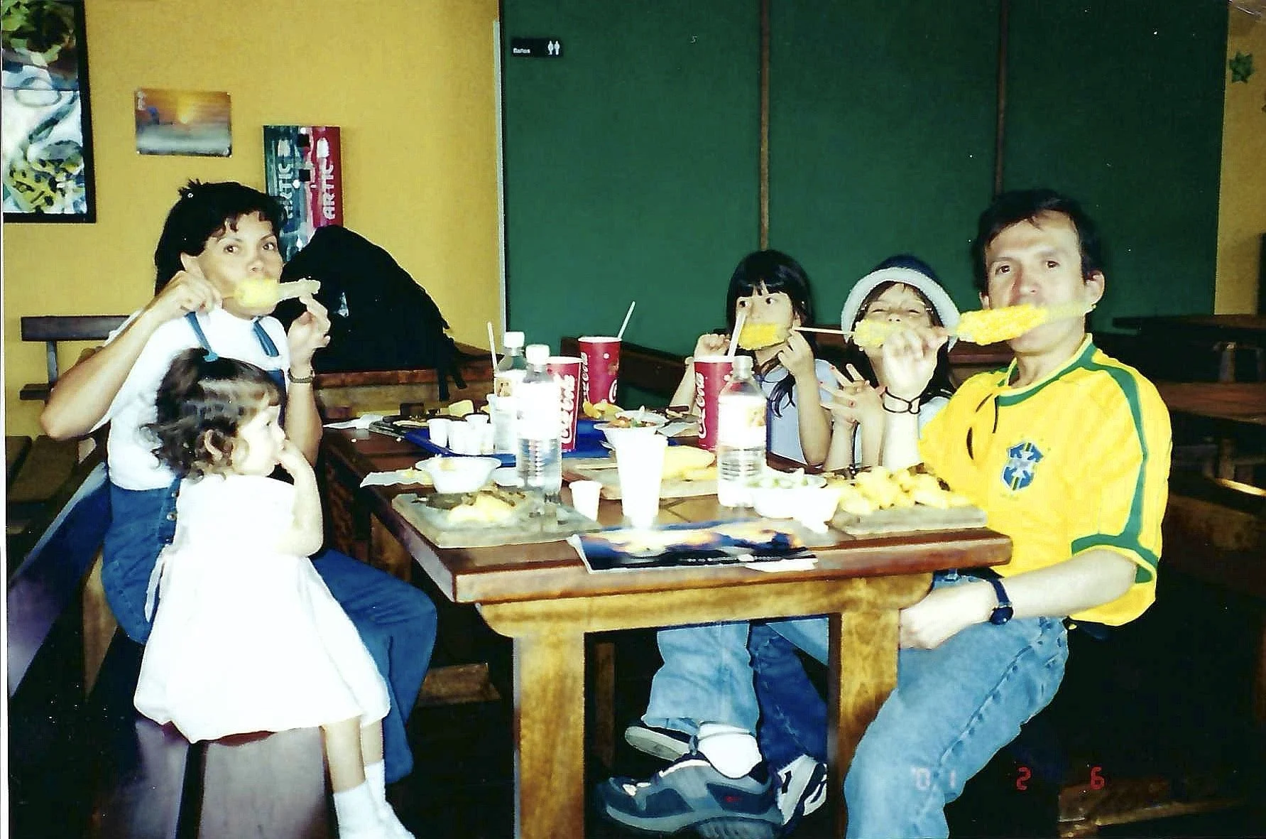A group of five people, two adults and three children, sit around a wooden table in a restaurant. They are enjoying a meal, with some of them eating food on skewers. The table has various drink bottles, cups, and food items. The background features a