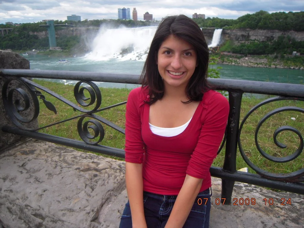 Smiling young woman with dark hair in a red top standing near a decorative metal railing at Niagara Falls, with the waterfall and city skyline in the background.