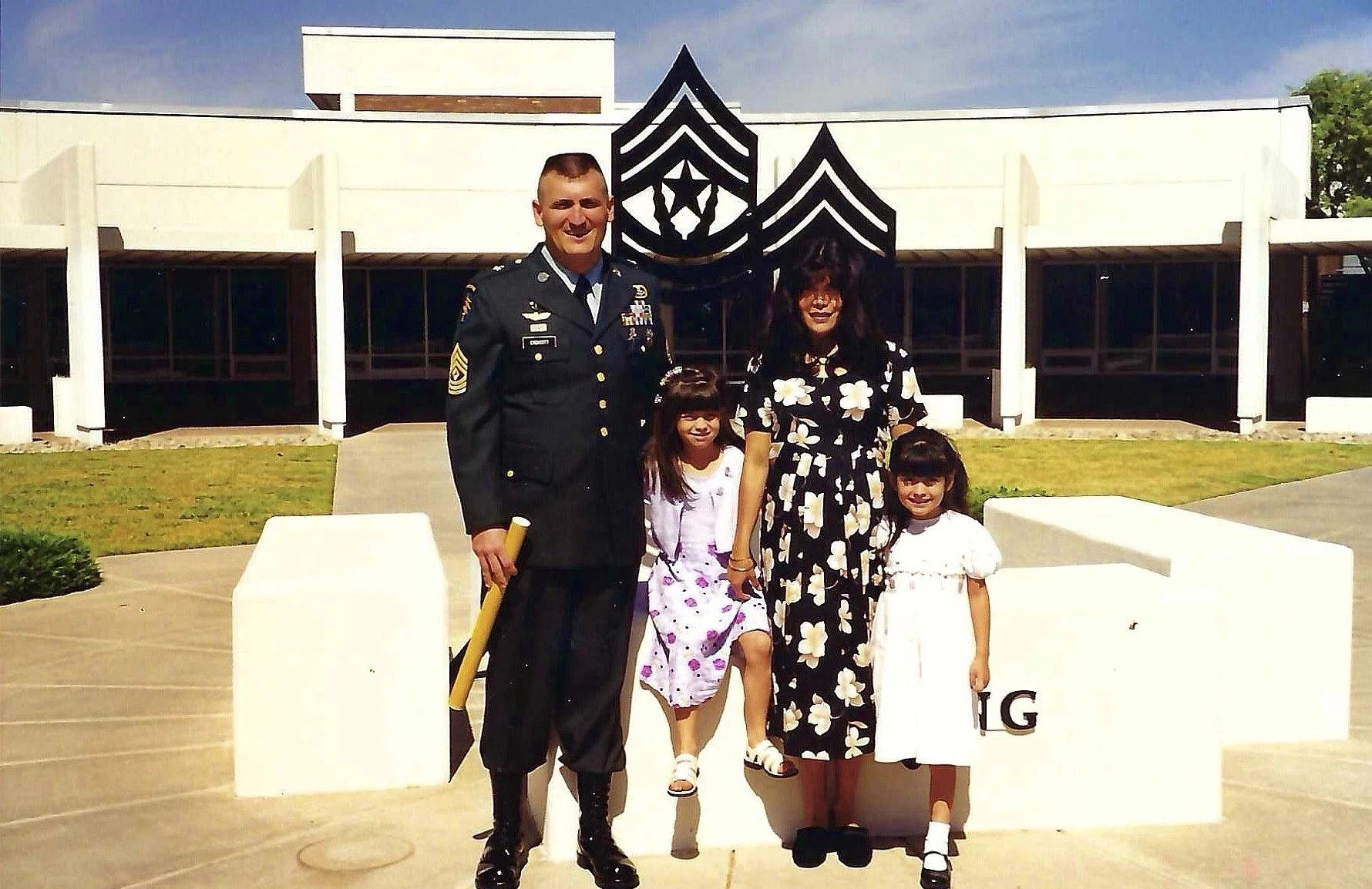 A man in a military uniform standing with two young girls and a woman, all posing together in front of a white monument with a black emblem and the letters IG on it, outdoors on a sunny day.