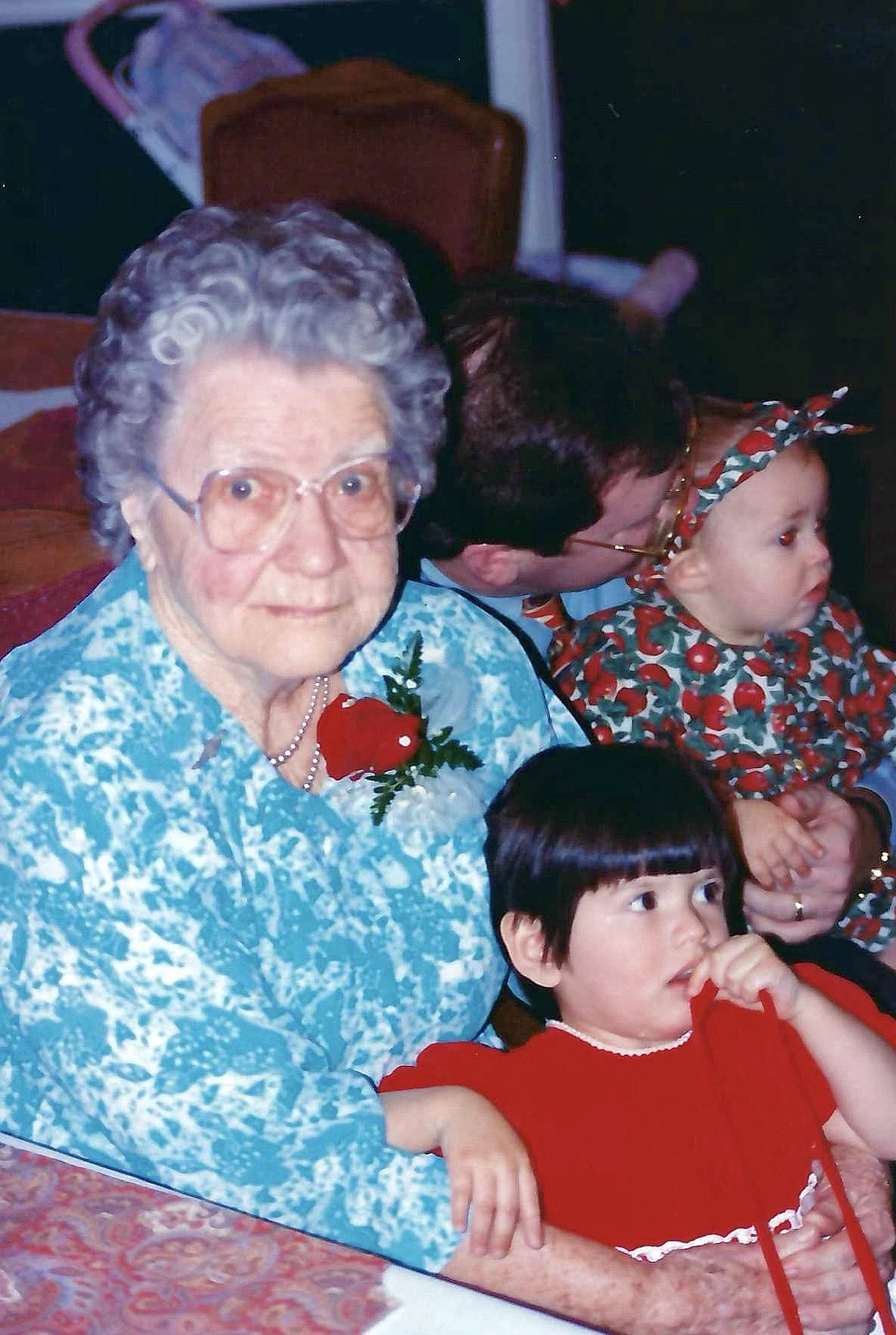 An elderly woman with gray curly hair, wearing glasses and a blue patterned dress, sits with three young children. One girl in a cherry-themed dress and matching headband, a boy in a red sweater, and another girl with dark hair partially visible. The