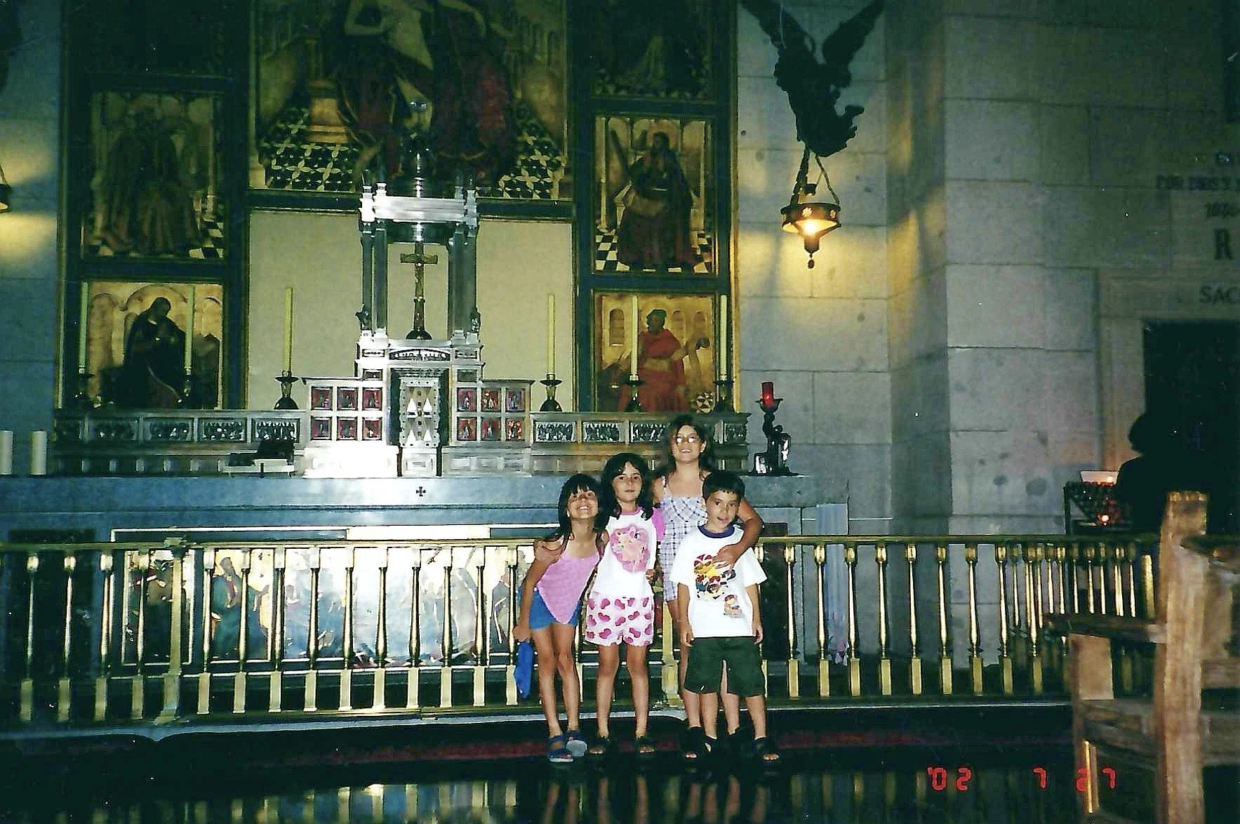 Four children standing in front of an altar inside a church, with religious artwork and statues in the background, and a statue of an angel hanging from the ceiling.