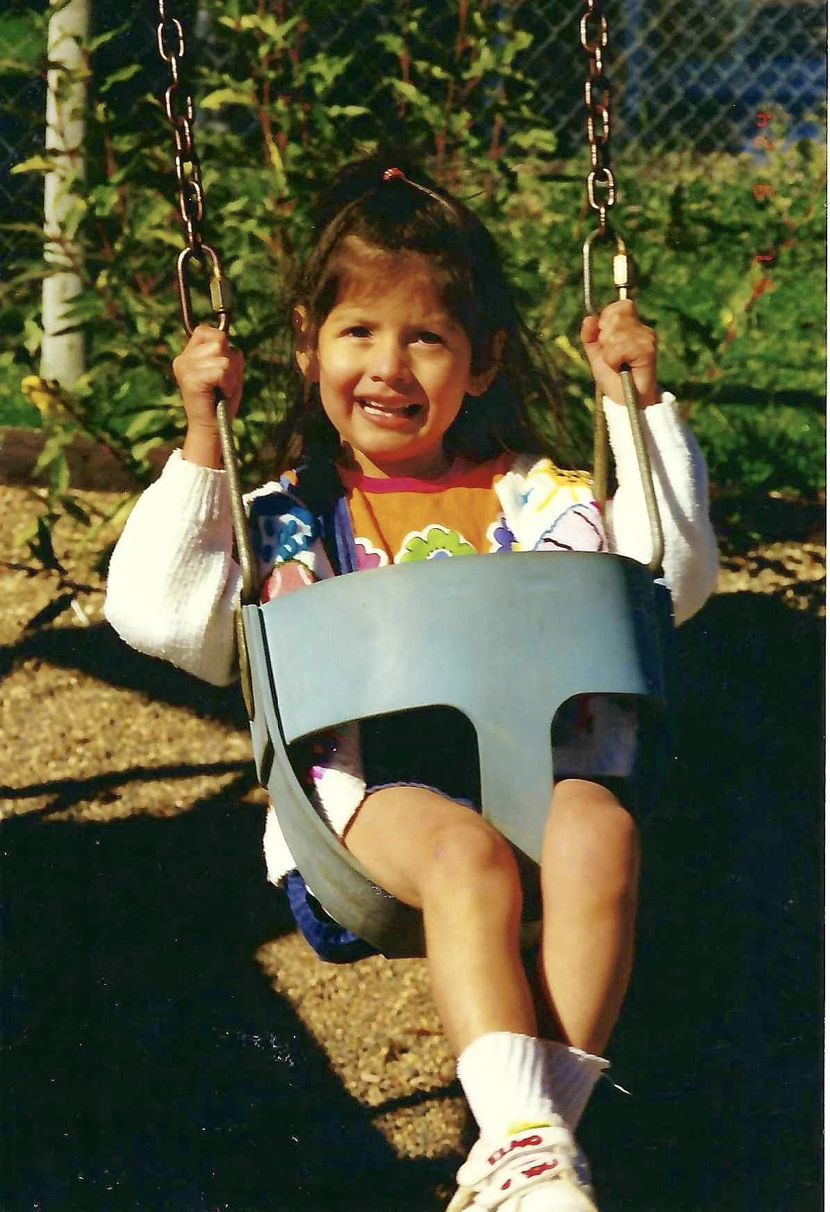 A young girl sitting on a swing at a playground, wearing a colorful shirt, white sweater, and white sneakers. She appears to be crying or upset.