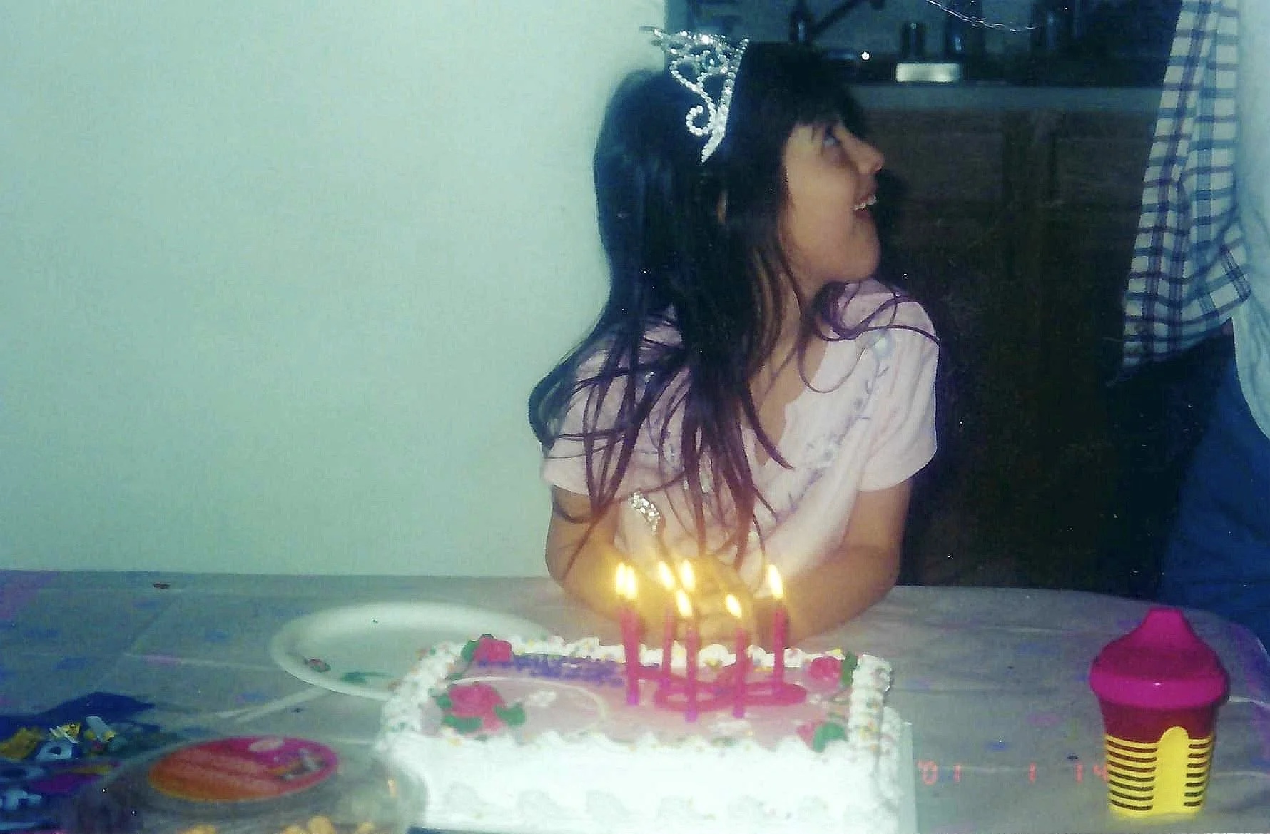 A young woman with a crown or tiara on her head celebrating a birthday, sitting at a table with a decorated birthday cake with pink candles, surrounded by colorful party items.