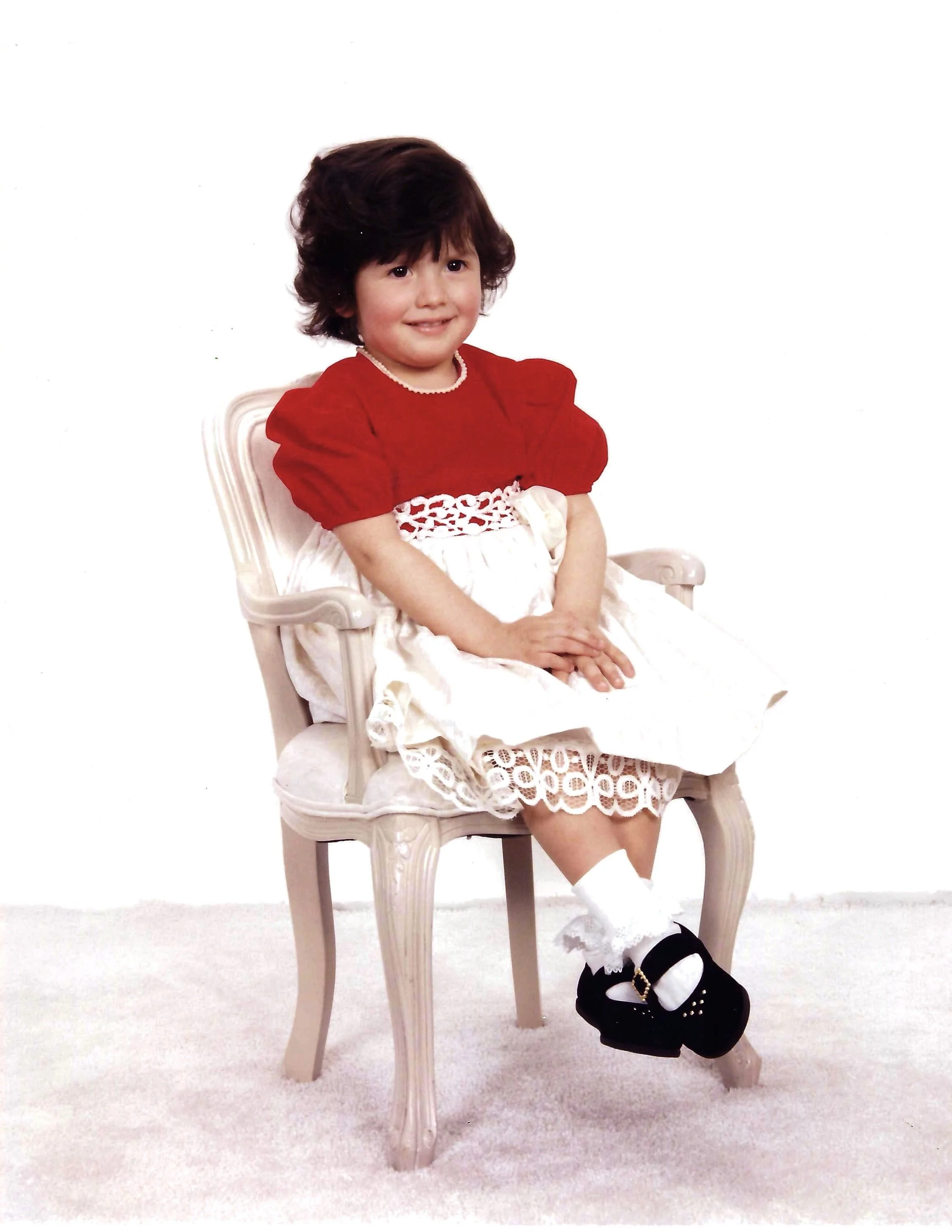 A young girl with dark hair smiling, wearing a red and white dress, sitting on a cream-colored chair with ornate legs, on a white carpeted floor.