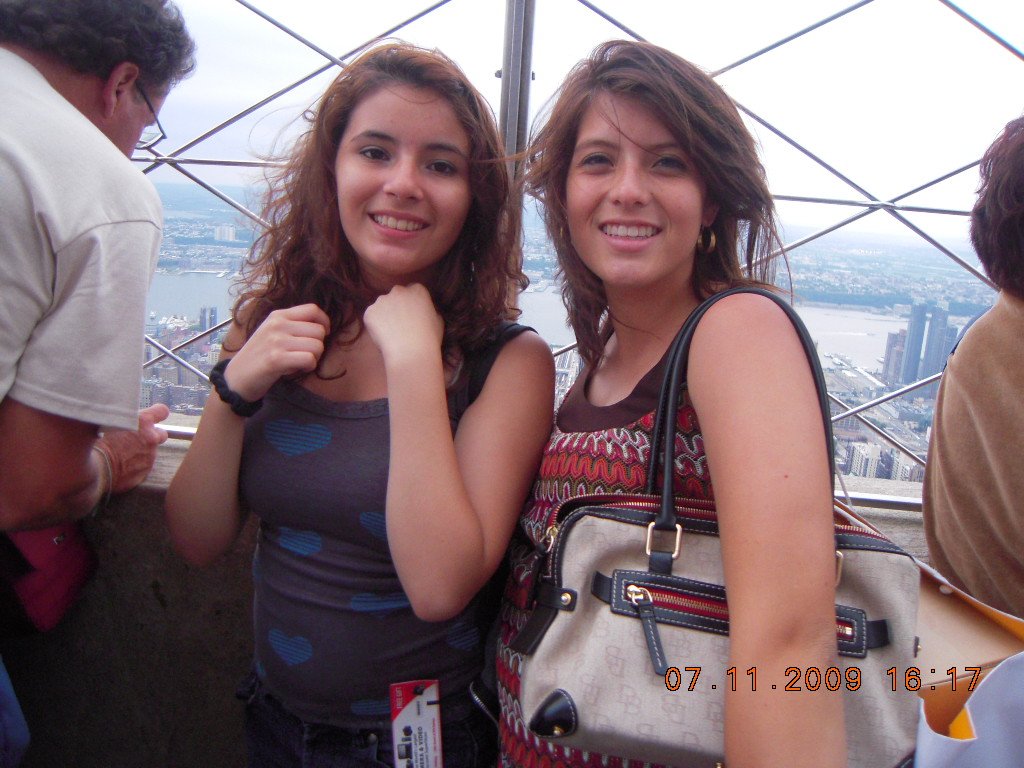 Two young women smiling on a viewing deck of a tall building with a city and river in the background, during daytime.