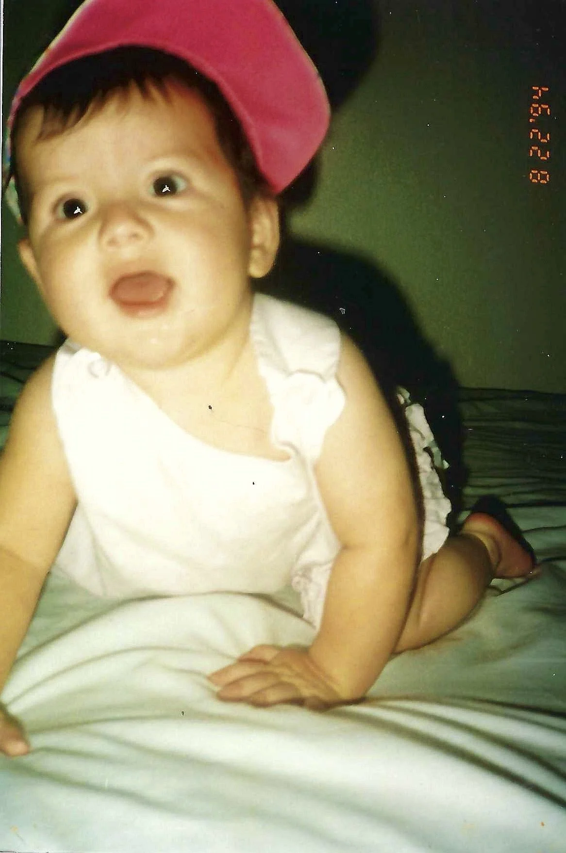 A young child with short dark hair, wearing a pink cap, a white sleeveless top, and sandals, lying on a bed with striped bedsheets, looking up and smiling.
