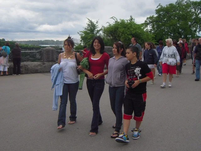 Group of four women and one boy walking outdoors near a railing with other people around and trees in the background.