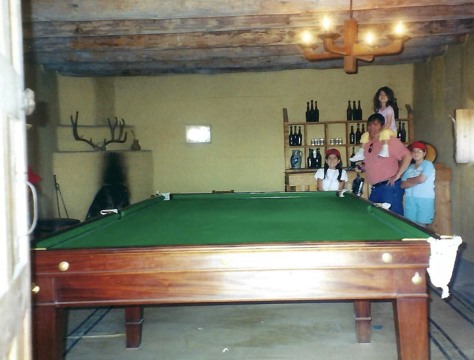 A game room with a pool table, four children, and a man. The children are wearing hats, and one of them is riding on the man's shoulders. The room has a wooden ceiling, a chandelier, and a shelf with bottles and jars.