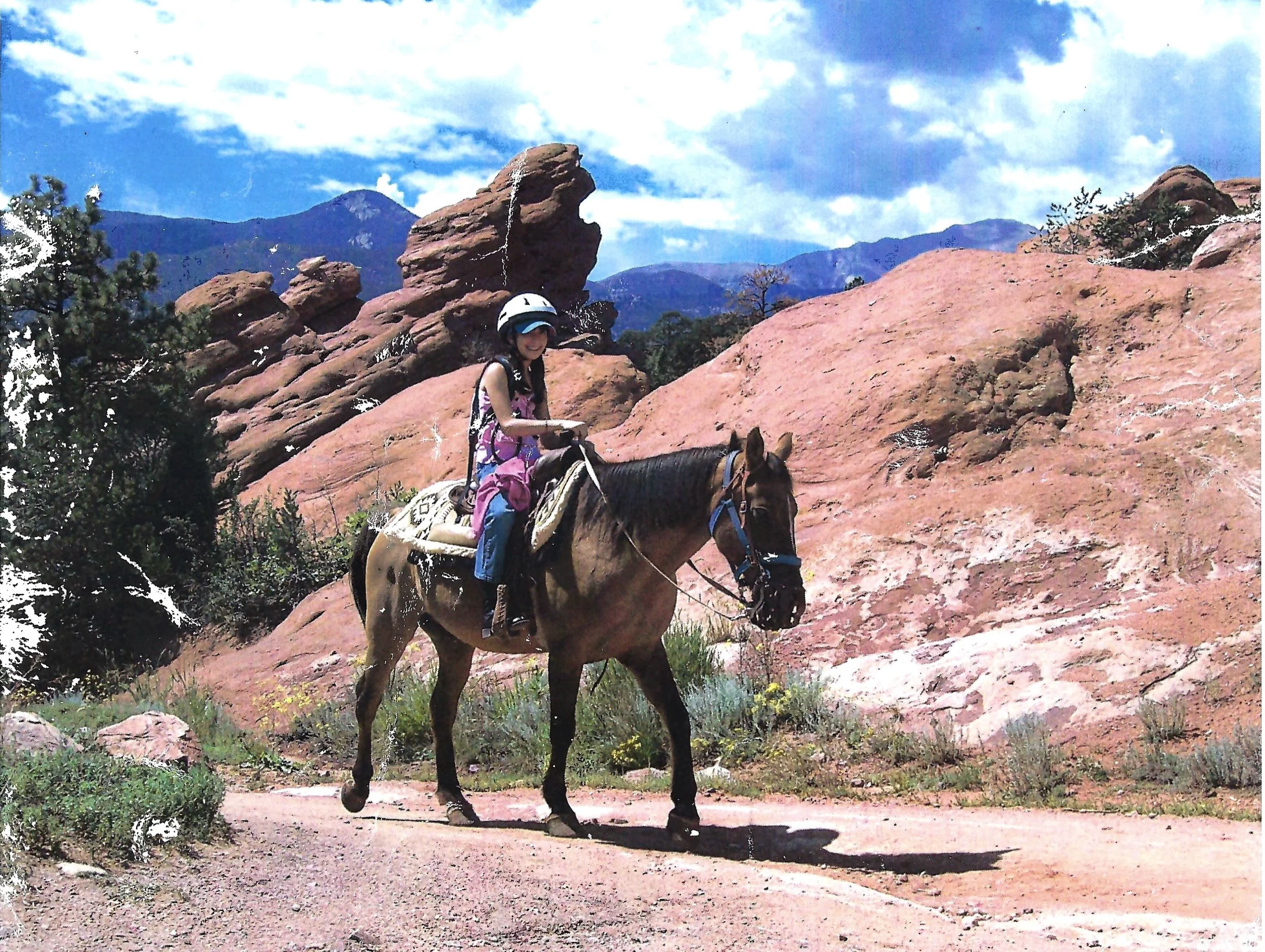 A person riding a horse across a rocky landscape with reddish rock formations, trees, and mountains in the background under a partly cloudy sky.