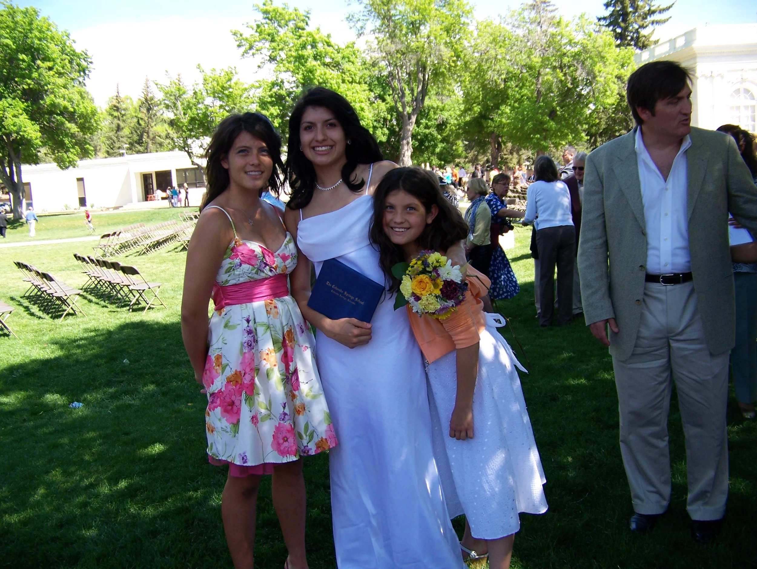 Three women in formal dresses at an outdoor event, with one woman holding a bouquet of flowers and a diploma, smiling for the photo, surrounded by other people and chairs on a grassy area with trees.