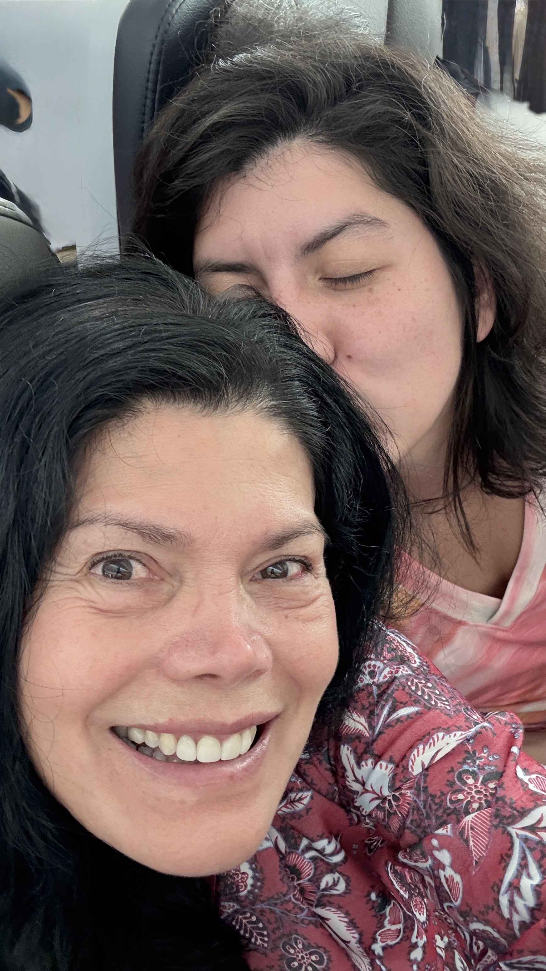 A woman smiling and a girl resting her head on her shoulder, sleeping, with the woman wearing a red floral shirt.