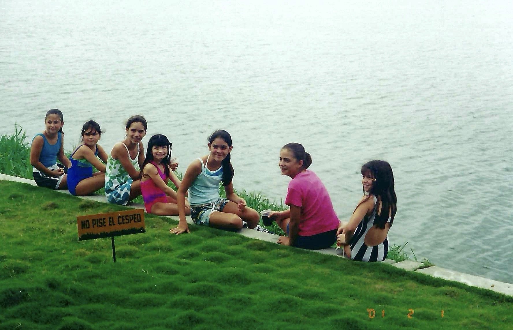 A group of children and two women sitting along the edge of a body of water, smiling and holding hands, wearing swimsuits and casual clothing, with a sign in Spanish that says 'No pise el cesped'.
