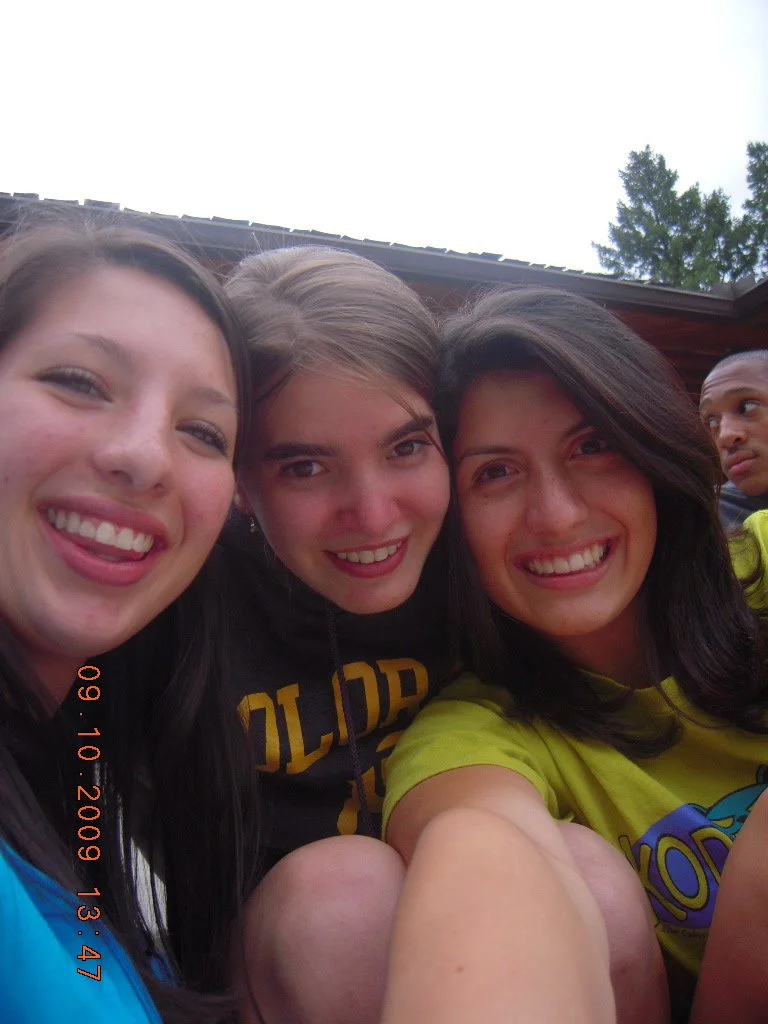 Three young women smiling and taking a close-up selfie outdoors, with another person partly visible in the background near a building and trees.