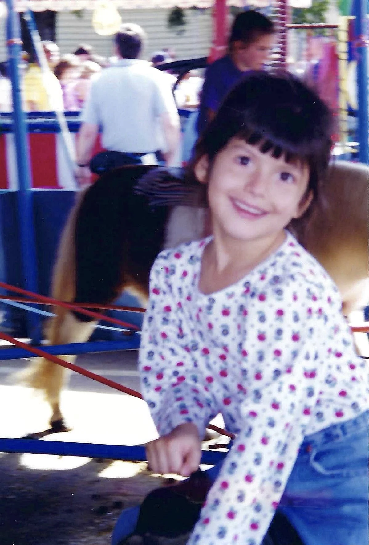 A young girl with short dark hair and a floral long-sleeve shirt riding a pony at an amusement park. She is smiling and holding the pony's rein. In the background, there are other park visitors and colorful structures.