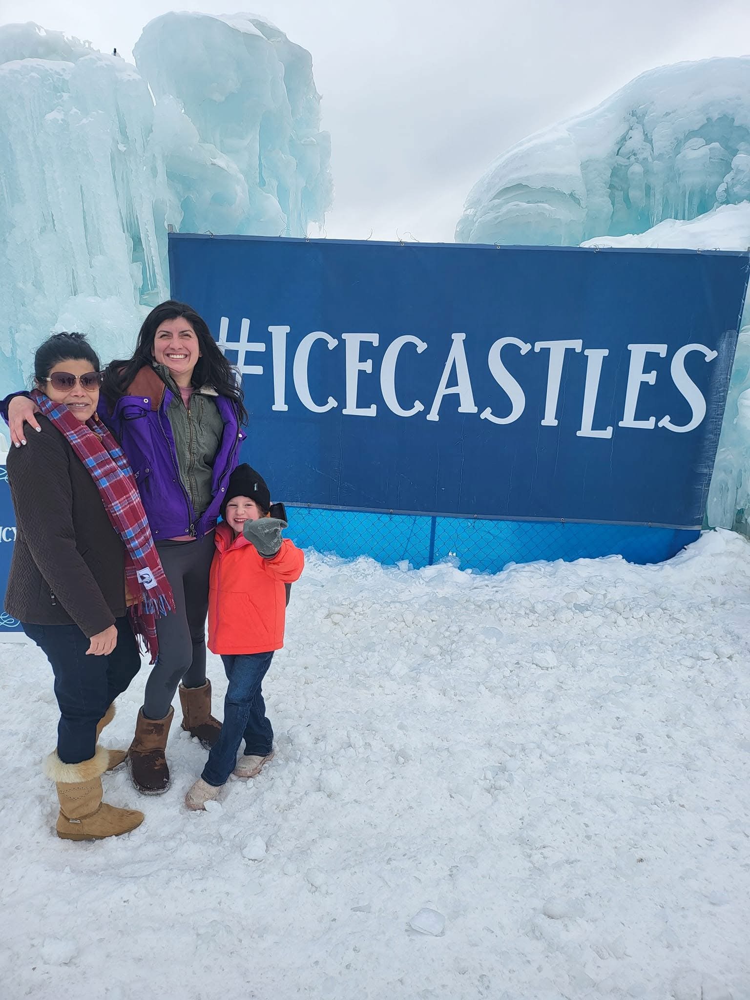 Group of three people, two women and a young girl, standing in the snow in front of a large blue sign with #ICECASTLES, with ice formations in the background.