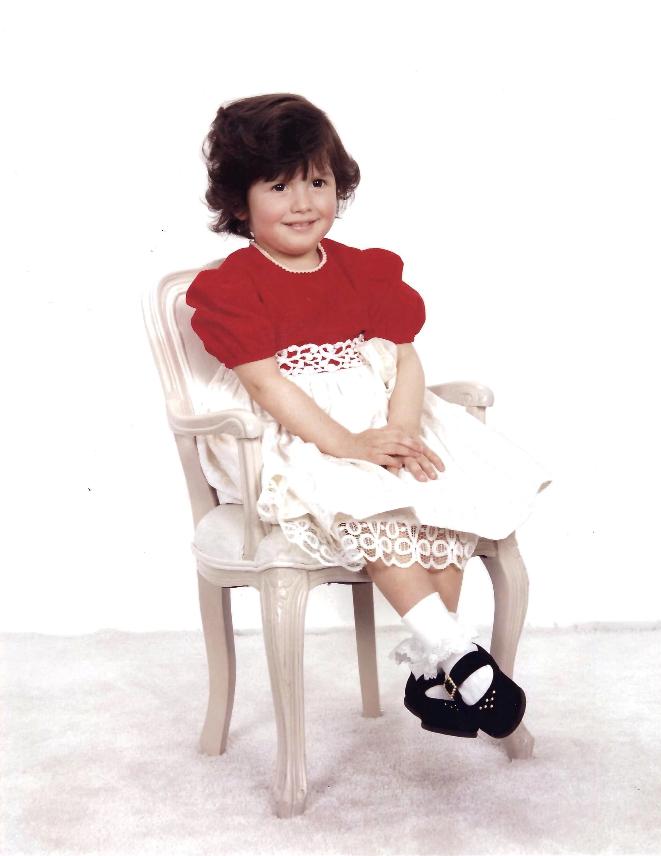 A young smiling girl with dark curly hair seated on a white vintage-style chair against a white background, wearing a red top, a white lace dress, and black shoes.