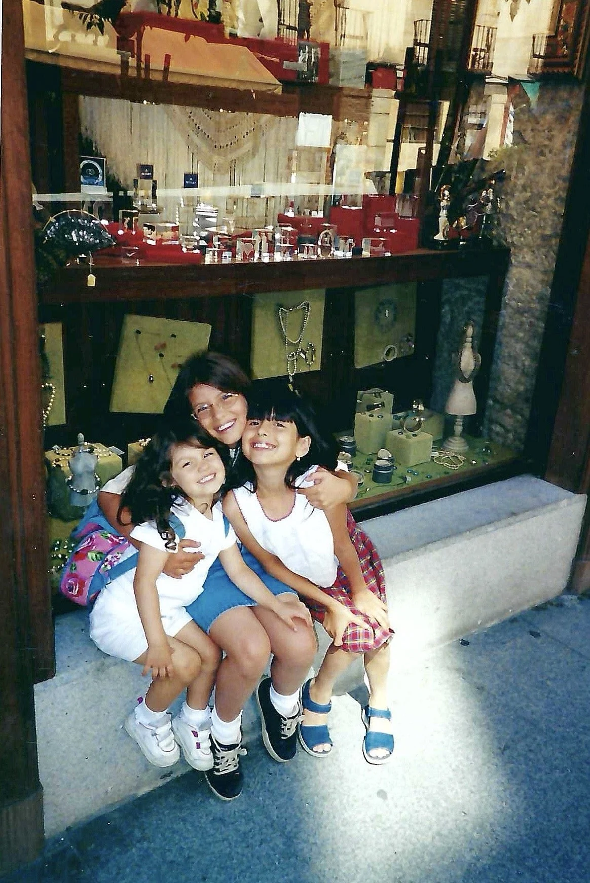 A woman and three young girls sitting on a stone bench in front of a large glass display case filled with jewelry, decorative items, and accessories. They are smiling and hugging each other.