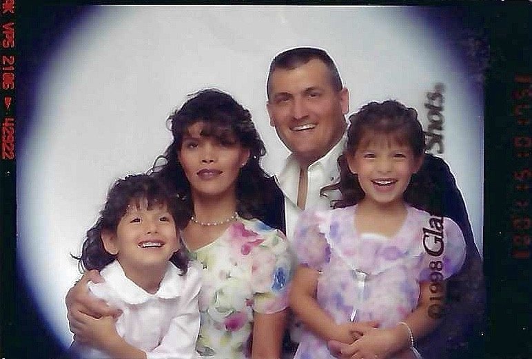 A family of four posing for a portrait, including a mother, father, and two young daughters, all smiling.