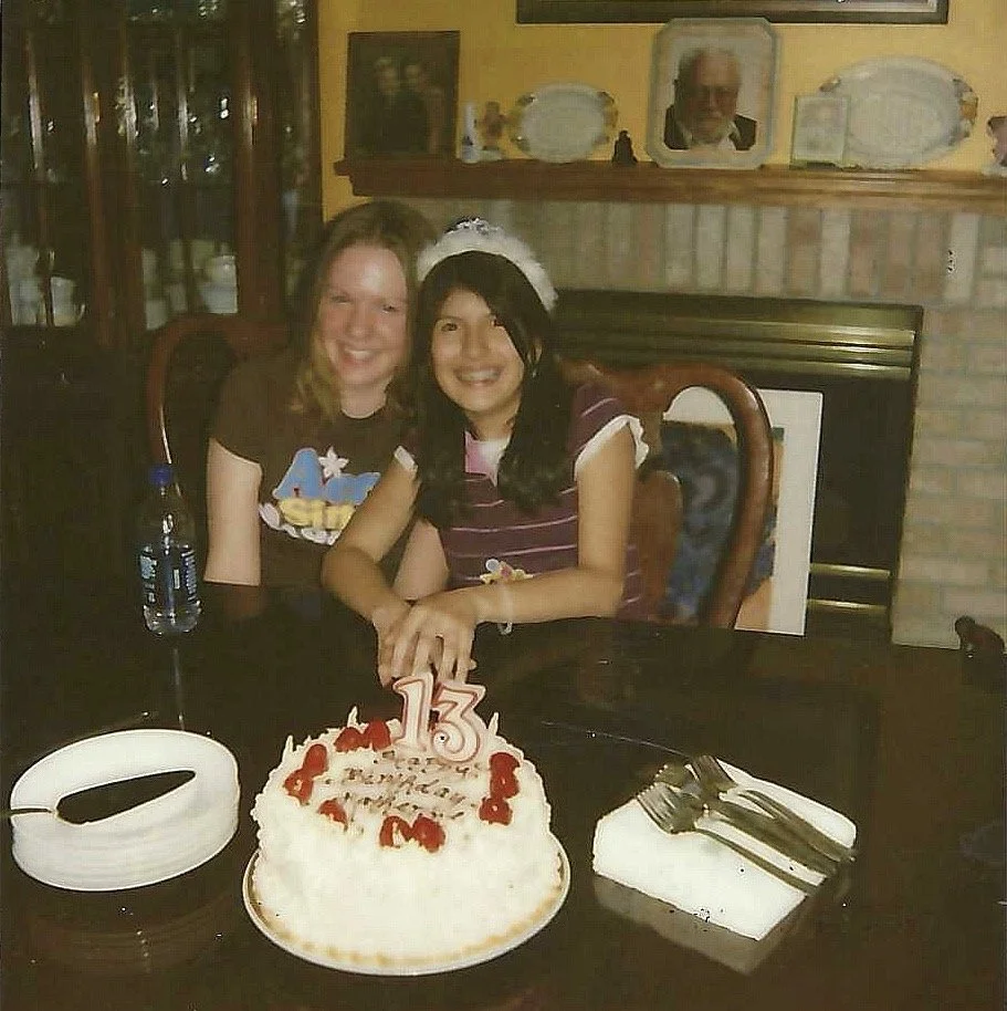 Two girls celebrating a birthday with a cake decorated with strawberries and a '13' candle, sitting at a table in a cozy living room.