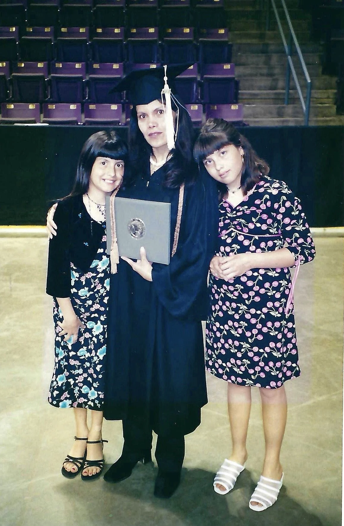 A woman in a graduation cap and gown holding a diploma, standing between two girls in floral dresses, at a graduation ceremony with empty purple seats in the background.