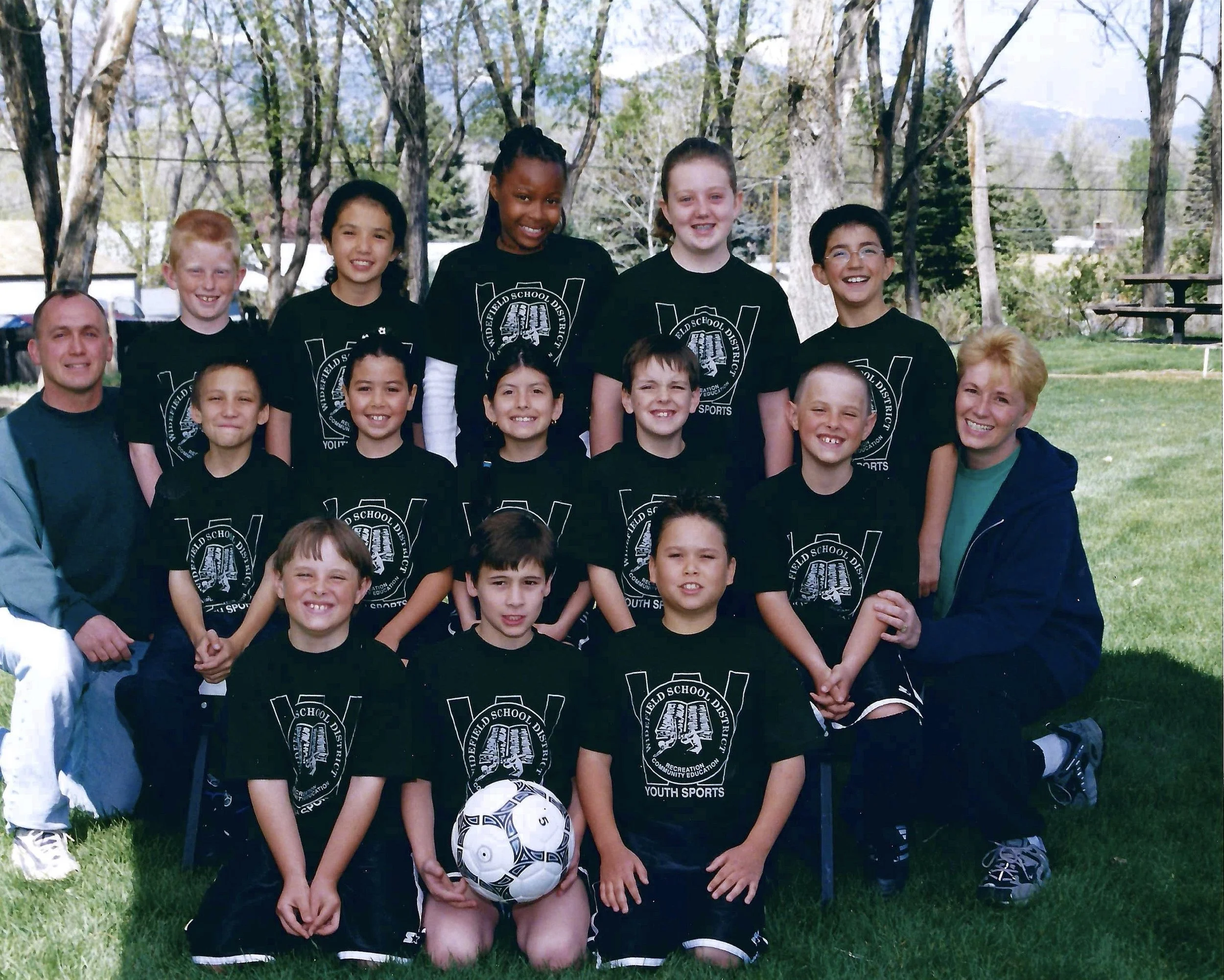 Group of children and two adults, standing outdoors on grass with trees in the background, wearing matching black T-shirts with school district logo and text related to youth sports or education.