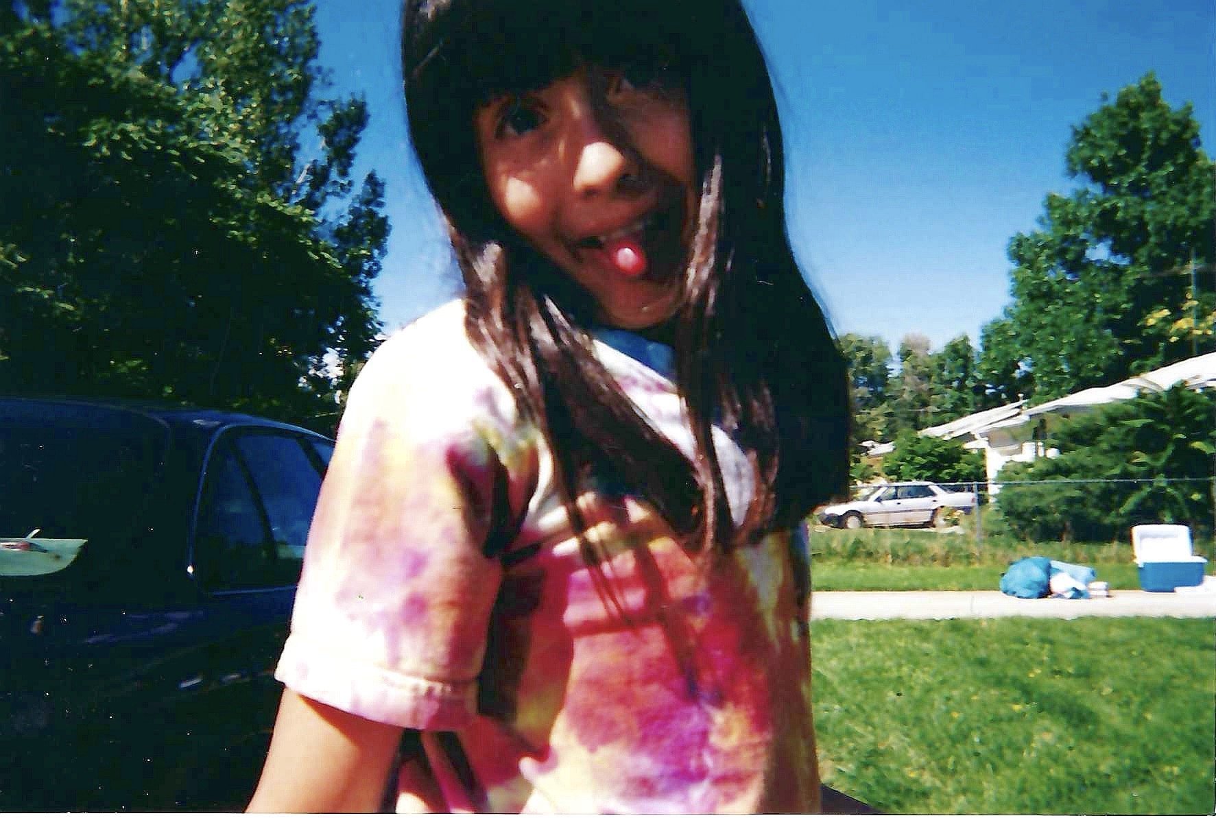 A young girl with dark hair, wearing a colorful tie-dye shirt, sticks out her tongue with a small pink object on it, smiling outdoors on a sunny day. In the background, there are trees, a blue sky, parked cars, and some items on the grass.