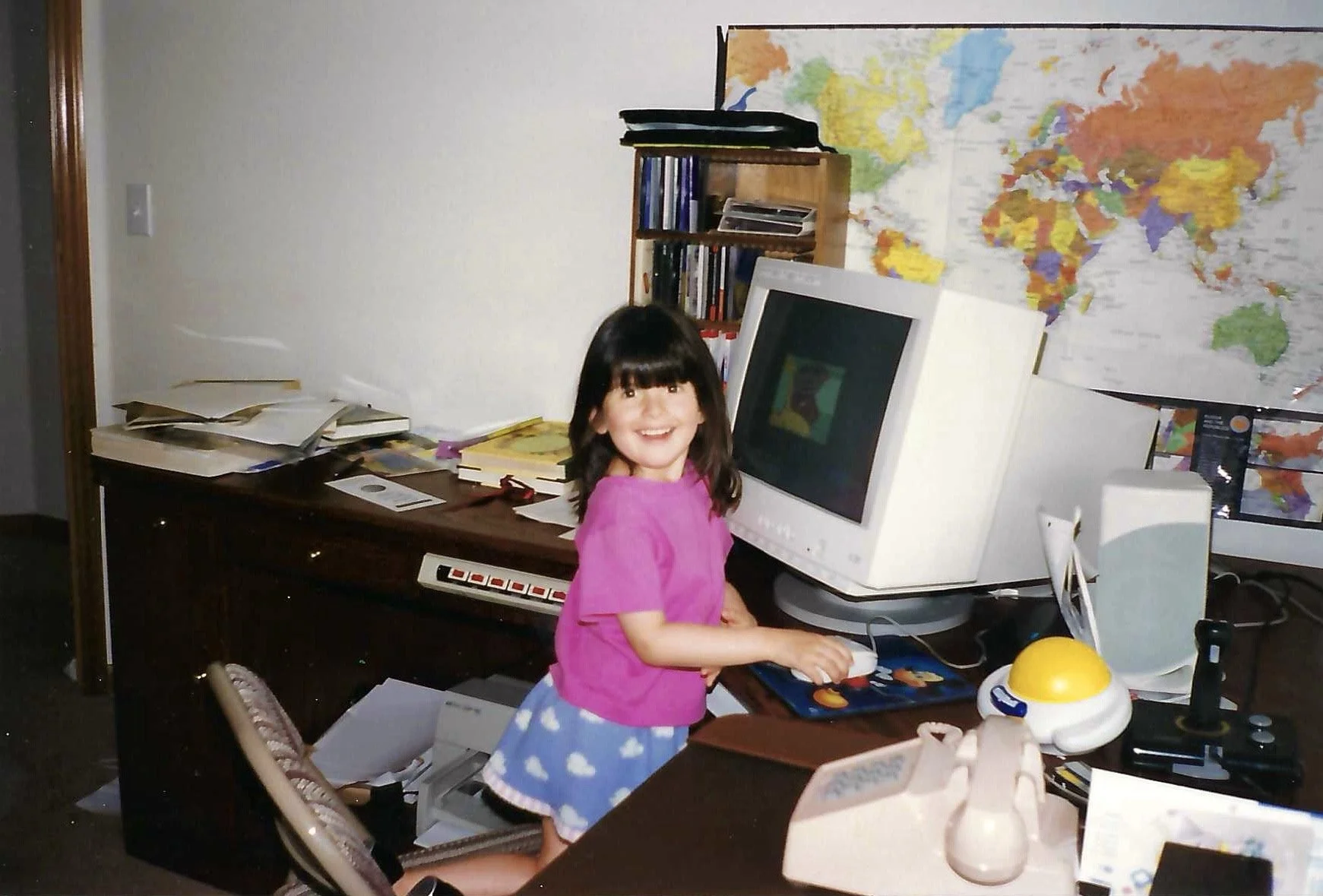 A young girl with dark hair and a bright smile working at a cluttered desk with a computer, in front of a world map on the wall.